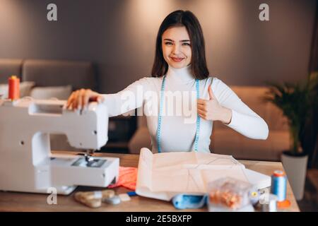 Young dressmaker woman sews clothes on sewing machine. Seamstress happy and show thumb up in workshop. Creating online clothing design courses. Stock Photo