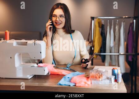 Young dressmaker woman sews clothes on sewing machine. Smiling and talking on phone seamstress in workshop. Creating online clothing design courses. Stock Photo