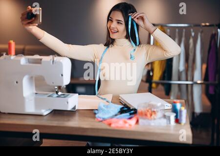Young dressmaker woman with sewing machine. Seamstress is taking selfie in workshop. Creating online clothing design courses. Stock Photo