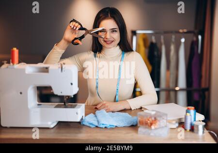 Young dressmaker woman sews clothes on sewing machine. Smiling and playing with scissors seamstress in workshop. Creating online clothing design Stock Photo
