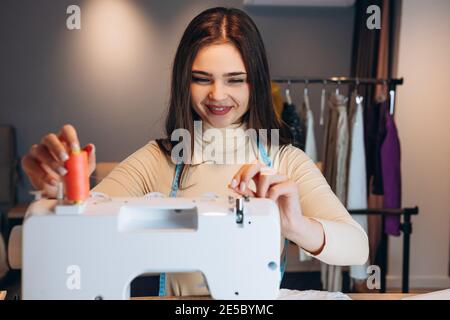 Young dressmaker woman sews clothes on sewing machine. Smiling seamstress in workshop. Creating online clothing design courses. Stock Photo