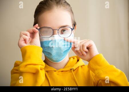 Foggy glasses wearing on young woman. Teenager girl in medical protective face mask and eyeglasses wipes blurred foggy misted glasses. New normal due Stock Photo