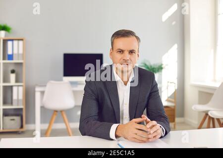 Serious confident businessman sitting at office desk, looking at camera and listening to you Stock Photo