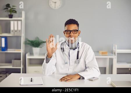 Online doctor sitting at desk and waving hand at camera, greeting patient during video call Stock Photo