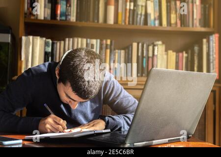 Young man taking notes in a notebook while working or studying online on a laptop with a bookcase in the background in concept of studying and working Stock Photo