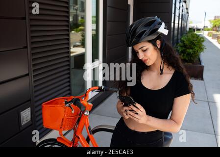 Woman wearing bike helmet looking at phone in front of orange bike Stock Photo