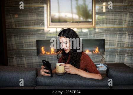 Happy young woman holding phone and drinking coffee on couch Stock Photo