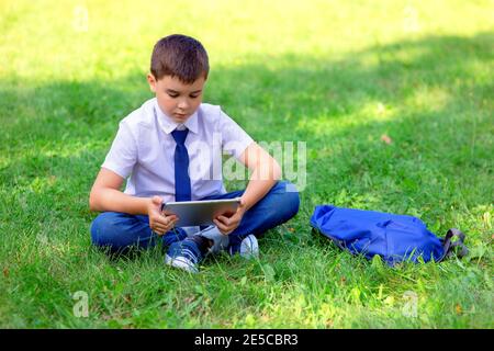 Serious Schoolboy in a white shirt and blue tie sits on green grass and plays with a tablet Stock Photo