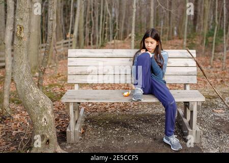 A tween girl sits alone on a bench in woods eating an orange in autumn Stock Photo