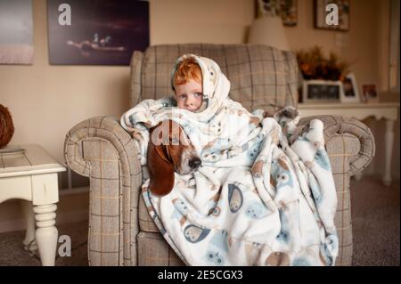 Boy 3-4 years old cuddles with dog in large blanket sitting in chair Stock Photo