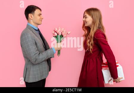 Beautiful romantic couple, attractive young woman in a red dress makes a gift for her man in a suit, on a pink background, Happy Valentine's Day Stock Photo