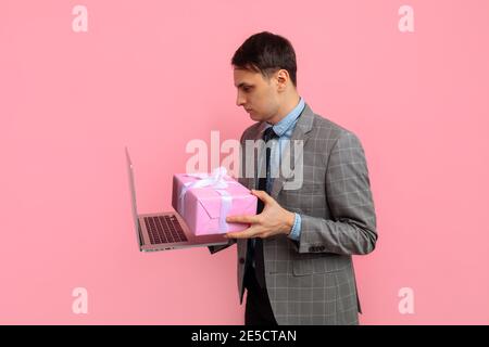 Elegant young man in a suit, with a gift box uses a laptop computer, on a pink background, the concept of Valentine's day, shopping Stock Photo
