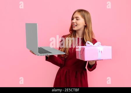 Happy attractive young woman in elegant red dress with gift box uses laptop computer, on pink background, concept of Valentine's day, shopping Stock Photo