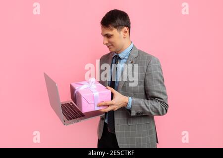 Elegant young man in a suit, with a gift box uses a laptop computer, on a pink background, the concept of Valentine's day, shopping Stock Photo