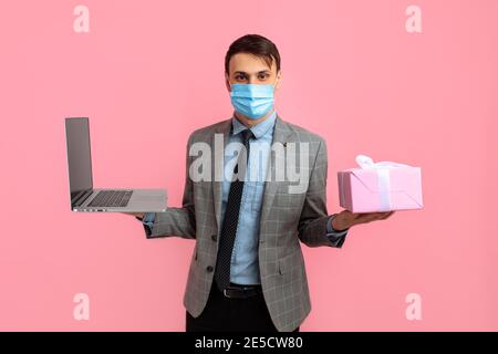 Elegant young man in a suit and medical mask, with a gift box uses a laptop computer, on a pink background, Valentine's day concept, shopping Stock Photo
