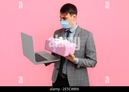 Elegant young man in a suit and medical mask, with a gift box uses a laptop computer, on a pink background, Valentine's day concept, shopping Stock Photo