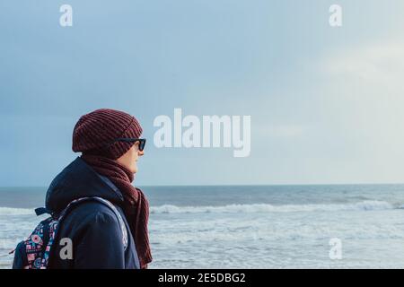 A young man in warm clothes and sunglasses walking on winter seaside and enjoying the moment. Relax during a walk on the coast. Local self-traveling