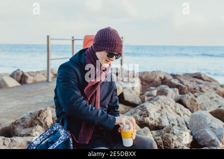 A young man in warm clothes and sunglasses drinking hot coffee or tea from reusable cup during walk on winter seaside. Relax on the coast. Local Stock Photo