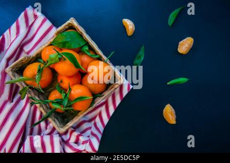 Top view Fresh ripe mandarins, clementine, tangerine with green leaves in the straw bowl on a striped napkin and dark blue stone background. Local Stock Photo