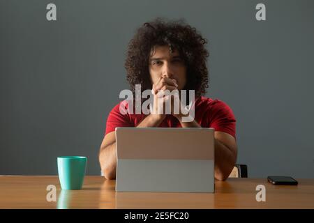 A CURLY HAIRED YOUNG MAN LOOKING AT CAMERA WHILE SITTING IN FRONT OF DIGITAL TABLET Stock Photo