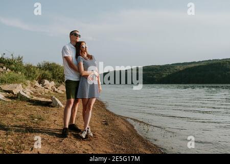 Portrait of couple in love standing on the beach at sunset. Full length of young woman and man hugging at the lake on warm summer evening. Stock Photo