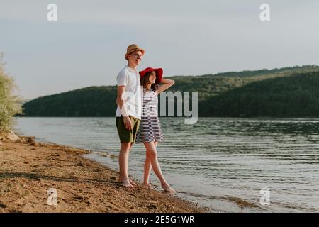 Full length of couple in love relaxing on the beach on lovely summer evening. Portrait of young woman and man hugging at the lake at sunset. Stock Photo