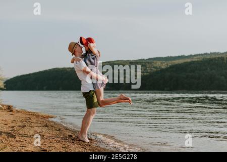Full length of happy couple in love having fun on the beach on lovely summer evening. Portrait of cheerful young man lifting his girlfriend at the lak Stock Photo