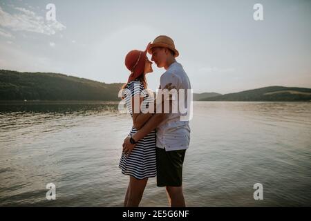 Couple in love hugging on the beach at sunset. Portrait of young man and woman looking into each other eyes at the lake against evening sun. Stock Photo