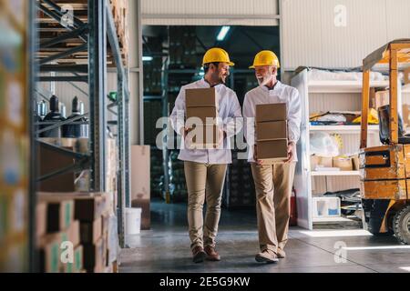 Picture of two male warehouse workers with helmets on their heads carrying boxes in their hands. Talking and walking. Stock Photo