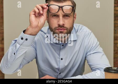 Handsome young man in a shirt sitting on the white background and frowning while looking from under the glasses Stock Photo