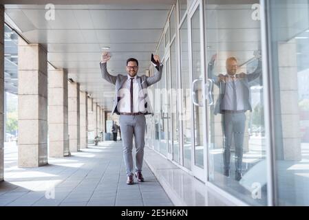 Happy businessman lifting arms while passing shop window. In one hand tablet and smart phone in other. Stock Photo