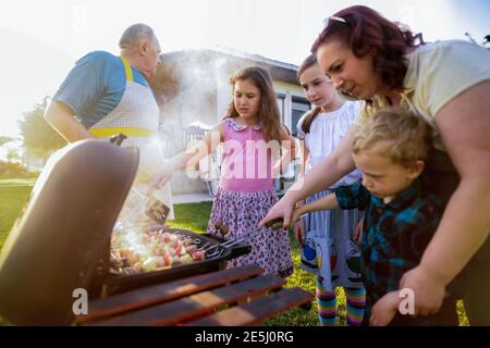 Cute little boy train to turn meat on barbeque grill.Other members of family standing around him and watching. Stock Photo