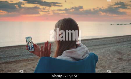 Young woman making video call with smartphone on the seashore. She speaking with young man. Stock Photo
