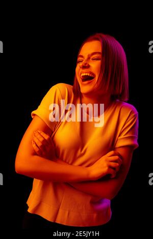 Crazy. Caucasian woman's portrait on black studio background in orange-pink neon light. Beautiful female model laughting delighted. Concept of human emotions, facial expression, sales, ad, fashion. Stock Photo