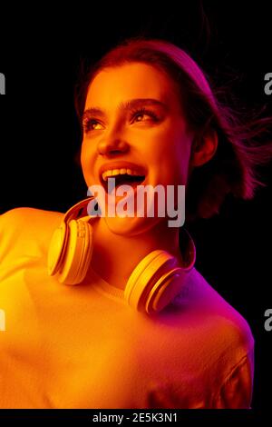 Happy, crazy. Caucasian woman's portrait on black studio background in pink-orange neon light. Beautiful female model with headphones. Concept of human emotions, facial expression, sales, ad, fashion. Stock Photo
