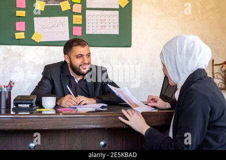 Handsome young businessman smiling and offering business contract to his pretty female Muslim business partner in white hijab for sign contract, sitti Stock Photo