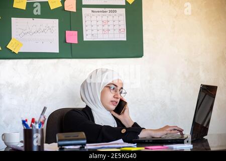 Muslim women working in the office from home using laptop and mobile phone for any support Stock Photo