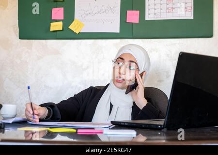 Muslim women working in the office from home using laptop and mobile phone for any support Stock Photo