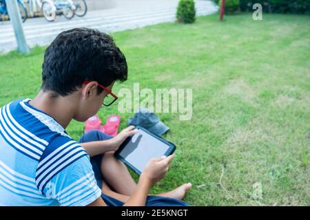 Teenager sitting on the lawn in the garden under the sun while using his tablet and wearing his glasses Stock Photo