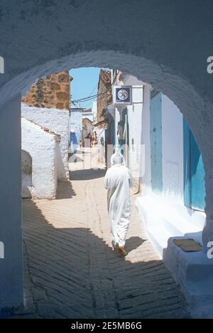 1984 Morocco Chefchaouen - Chefchaouen, Shafshāwan, also Chaouen, is a city in northwest Morocco. It is noted for its buildings in shades of blue. Chefchaouen is situated just inland from Tangier. The beauty of Chefchaouen's mountainous surroundings are enhanced by the contrast of the brightly blue painted medina (old town). People in traditional costume walking through the narrow blue painted streets of the Old town of Chefchaouen Morocco North Africa Stock Photo