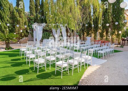 Outdoors wedding ceremony setup with white chairs in a garden with trees in the background. Stock Photo