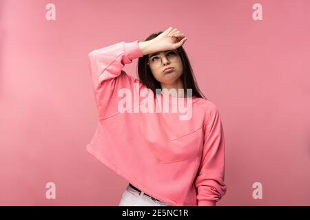 Photo of attractive young woman in glasses looks tired and exhausted, wears casual pink t-shirt white pants isolated pink color background Stock Photo