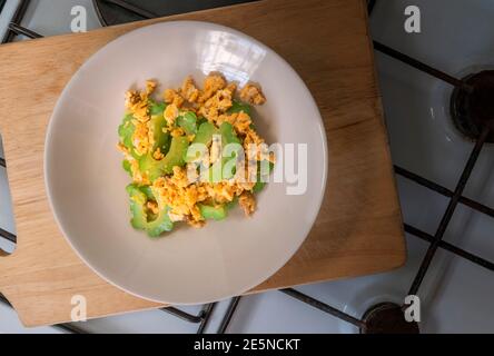Stir-fried bitter gourd with egg in round dish. Flat lay healthy food dish on wooden cutting board which put on stove as background. Top view food ima