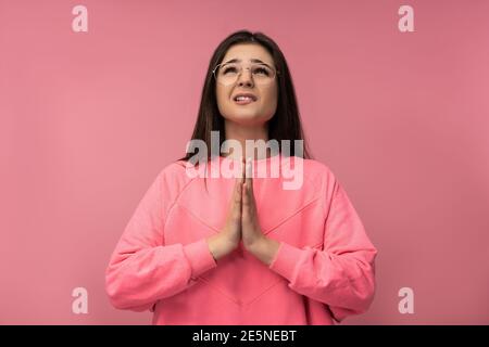 Photo of attractive young woman in glasses uncertainly praying , wears casual pink t-shirt isolated pink color background Stock Photo