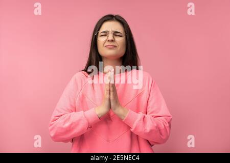 Photo of attractive young woman in glasses praying strongly, asks for something. Wears casual pink t-shirt isolated pink color background Stock Photo