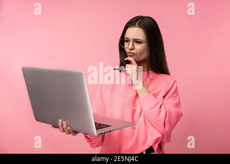 Photo of attractive lady in glasses interested and making decision, holds laptop and works online. wear casual pink t-shirt white pants isolated pink Stock Photo