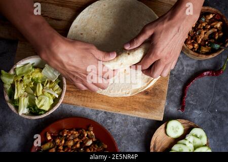 a young caucasian man, seen from above, rolls a durum or a burrito, filled with chicken meat cooked with different vegetables such as onion or red and Stock Photo