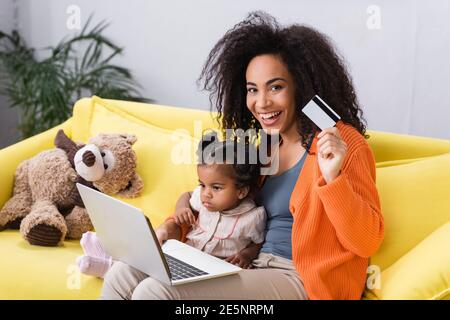 smiling african american mother holding credit card and using laptop near toddler daughter and soft toy Stock Photo