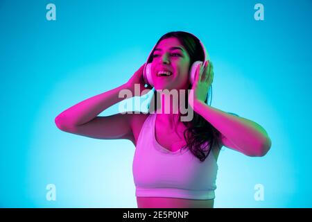 Joyful. Brunette woman's portrait on blue studio background in mixed neon. Beautiful model with headphones listens to music. Concept of human emotions, facial expression, sales, ad, fashion. Stock Photo