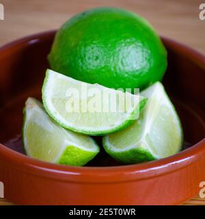 Whole green lime with cut pieces of lime in a brown ceramic dish and selective focus on a wood background Stock Photo